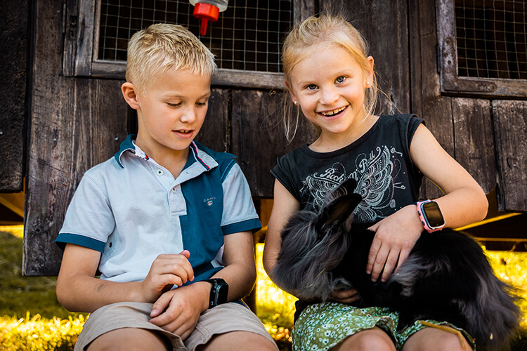 Zwei Kinder sitzen vor einer Holztür, eines hält ein schwarzes Kaninchen auf dem Schoß