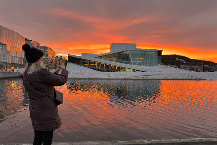 Person mit Winterjacke und Mütze fotografiert die moderne Oper in Oslo bei Sonnenuntergang am Wasser