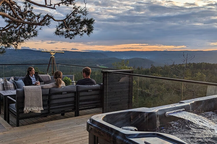 Drei Personen sitzen auf einer Terrasse mit Glasgeländer und Blick auf bewaldete Hügel bei Abenddämmerung, im Vordergrund ein Whirlpool
