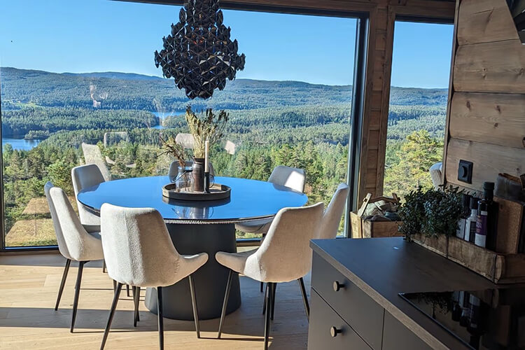 Runder Esstisch mit sechs gepolsterten Stühlen vor bodentiefen Fenstern mit Blick auf bewaldete Hügel und einen See