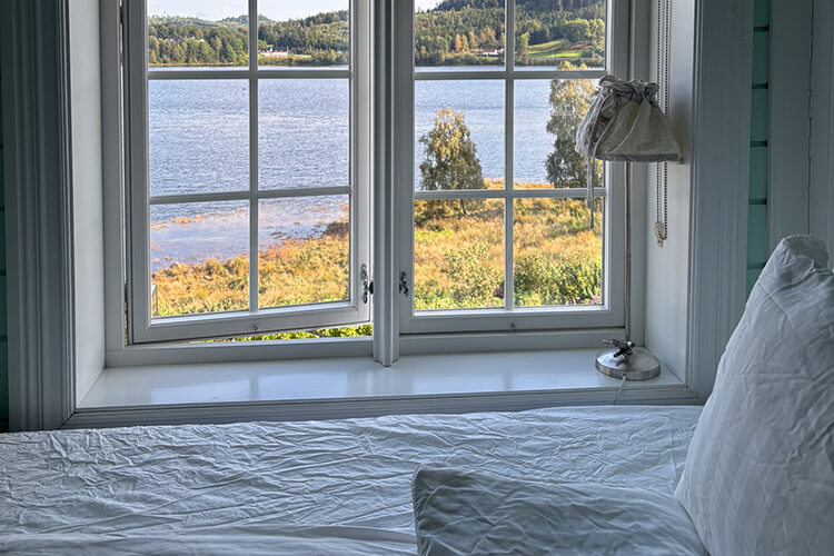 Ein Bett, dass direkt an einem Fenster steht. Durch das Fenster hat man einen Blick auf eine bewaldete Landschaft und einen großen Fluss.