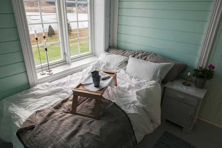 Ein Bett vor einem großen Fenster, mit Blick auf einen Fluss. Auf dem Bett steht ein Tablett mit Tassen.
