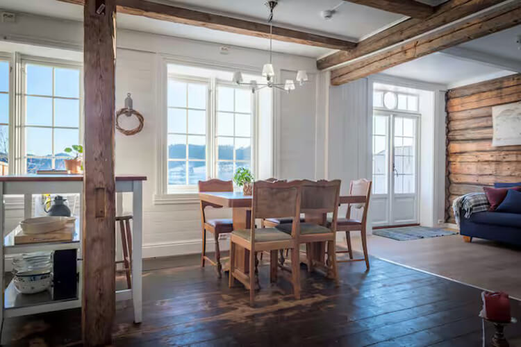 Ein Zimmer mit Holzboden und Holzbalken an den Wänden. Ein Esstisch steht im Zimmer und durch große Fenster sieht man auf einen Fluss.
