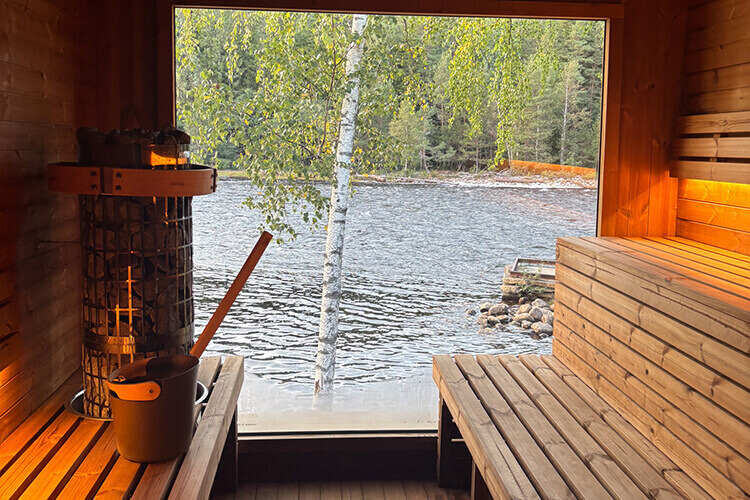 Eine Sauna mit einem großen Panoramafenster. Die Aussicht zeigt einen Fluss und eine Birke.