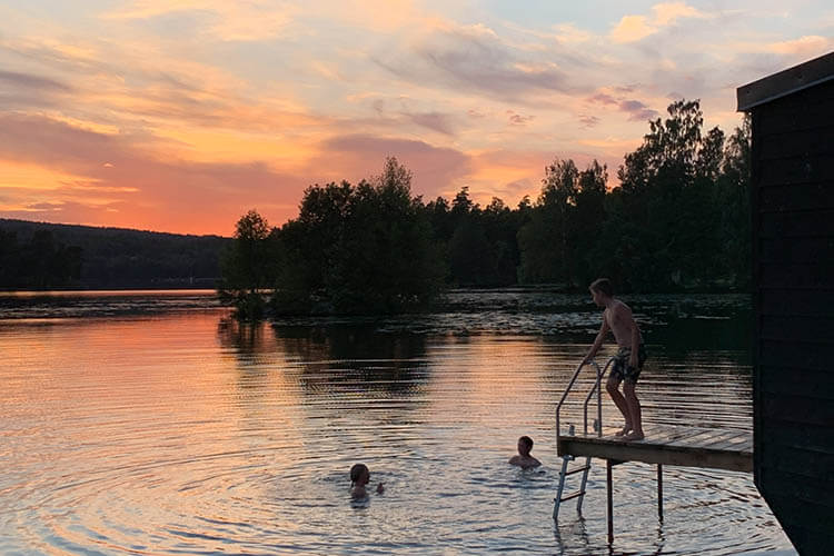 Drei Personen im See bei Sonnenuntergang, eine steht auf einem Steg mit Leiter an einer Sauna, zwei schwimmen im Wasser.
