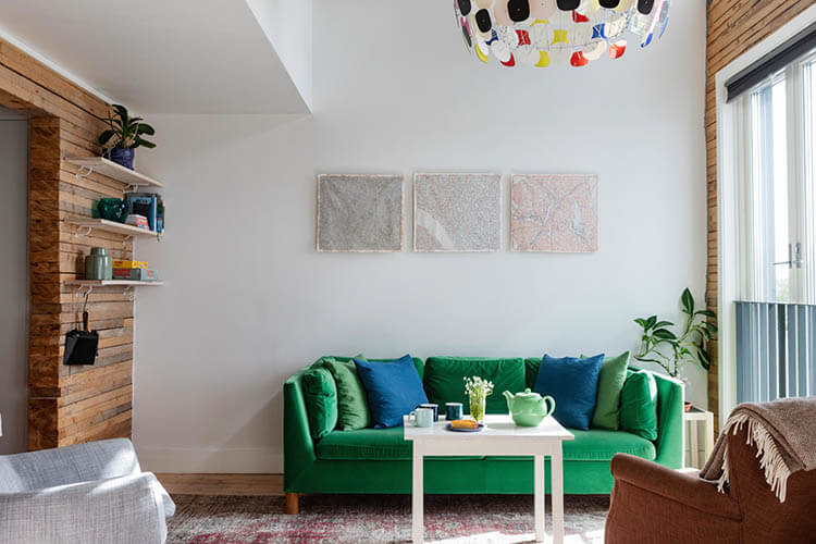 Hotelzimmer mit grünem Sofa, weißem Tisch, buntem Teppich und drei abstrakten Bildern an der Wand