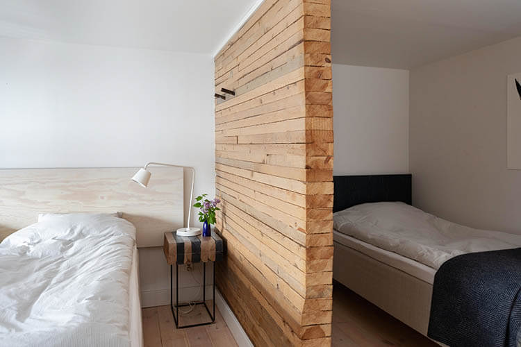 Zwei Schlafzimmer mit weißen Betten, getrennt durch eine Wand aus horizontalem Holz, mit einem kleinen Beistelltisch und einer weißen Lampe im Vordergrund