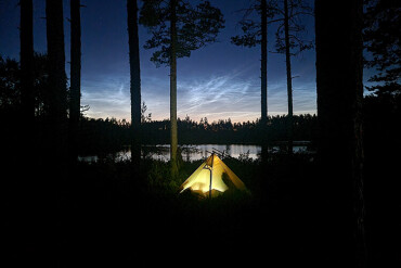 Beleuchtetes Zelt in einem Wald am Ufer eines Sees bei Nacht mit hellem Nachthimmel