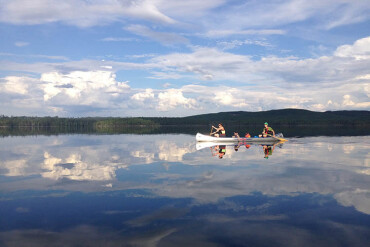 Drei Personen paddeln in einem weißen Kanu auf einem spiegelglatten See mit bewölktem Himmel und bewaldeten Hügeln im Hintergrund