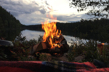 Lagerfeuer an einem Seeufer am Abend. Im Hintergrund Wald.