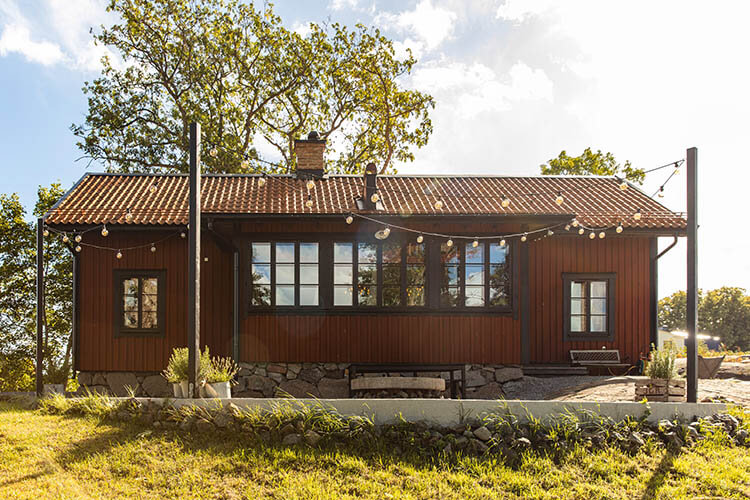 Ein eingeschossiges rotes Holzhaus mit Satteldach, umgeben von Bäumen und einer Steinmauer im Vordergrund