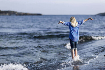 Frau mit blonden Haaren und blauem Hemd steht mit ausgebreiteten Armen im flachen Wasser an einer Küste