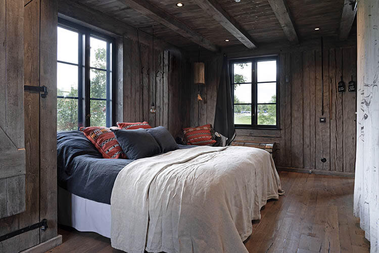 Rustikales Schlafzimmer mit Holzwänden, zwei Fenstern, einem großen Bett mit dunkler Bettwäsche und hellen Decken sowie bunten Kissen