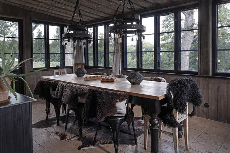 Rustikaler Holztisch mit mehreren Stühlen, die mit Fellüberwürfen bedeckt sind, in einem verglasten Raum mit Blick ins Grüne