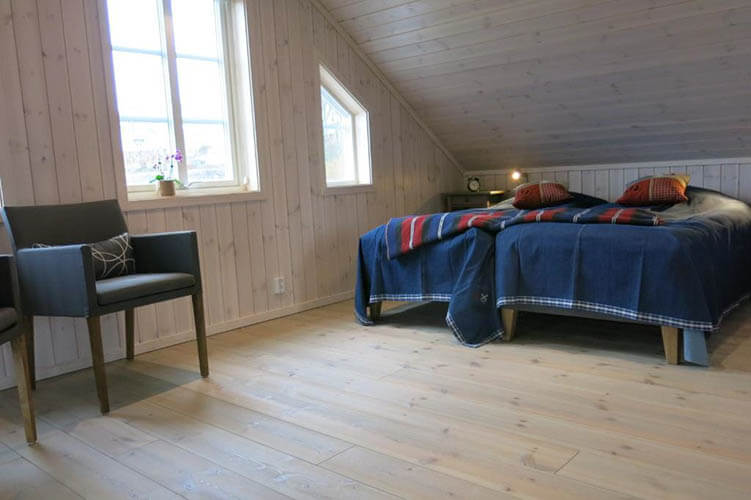 Schlafzimmer mit Dachschräge, zwei Betten mit blauen Decken und Kissen, grauer Sessel neben Fenster mit Pflanzen auf der Fensterbank