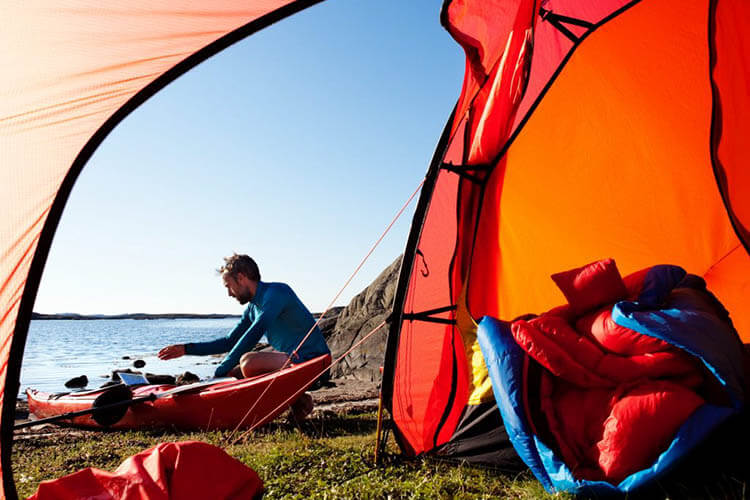 Person sitzt in einem Kajak am Ufer neben einem orangefarbenen Zelt mit Schlafsack und Ausrüstung