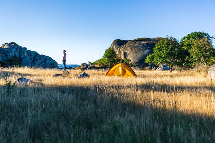 Zelt auf einer Wiese vor Felsen und Büschen, daneben stehende Person mit Blick in die Ferne