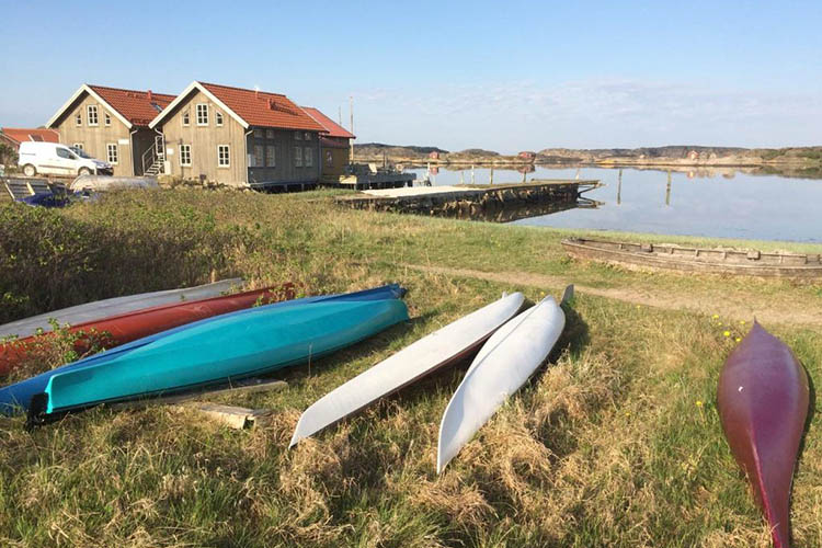 Mehrere Kanus liegen auf einer Wiese vor Holzhäusern an einem ruhigen Ufer mit Steg und weiter Landschaft im Hintergrund