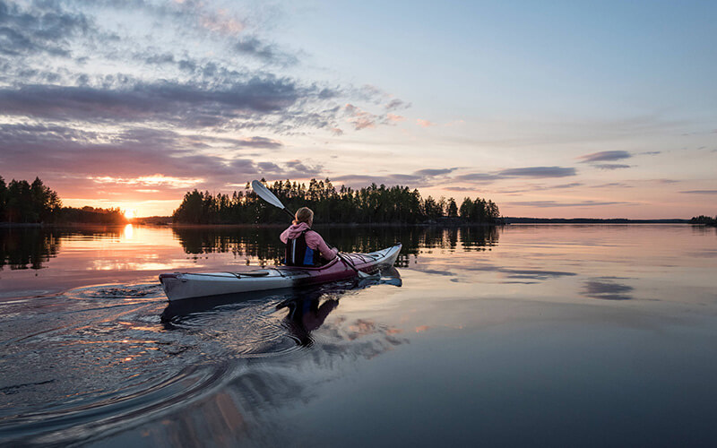 Person in Kajak auf ruhigem See bei Sonnenuntergang mit bewölktem Himmel und Bauminsel im Hintergrund.