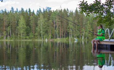 Kind mit grünem T-Shirt und Shorts sitzt auf einem Steg und angelt an einem ruhigen See mit Wald im Hintergrund.