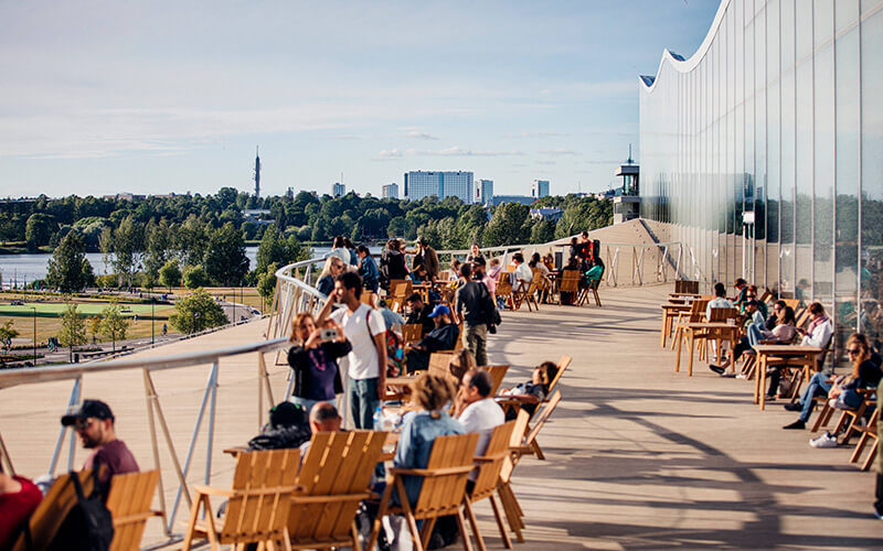 Menschen sitzen und stehen auf einer großen Holzterrasse mit Tischen und Stühlen neben einem modernen Gebäude mit Glasfassade und Blick aufs Meer und eine Stadt im Hintergrund.