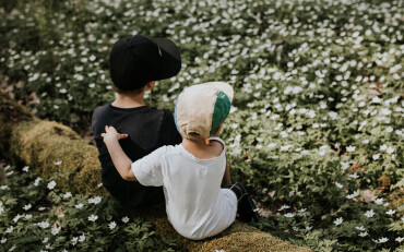 Zwei Jungen sitzen auf einem mit Moos bedeckten Baumstamm in einem blühenden Wald voller weißer Blumen.