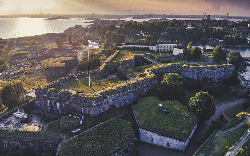 Luftaufnahme einer historischen Festungsanlage mit grasbewachsenen Mauern und einer finnischen Flagge auf einem Fahnenmast.