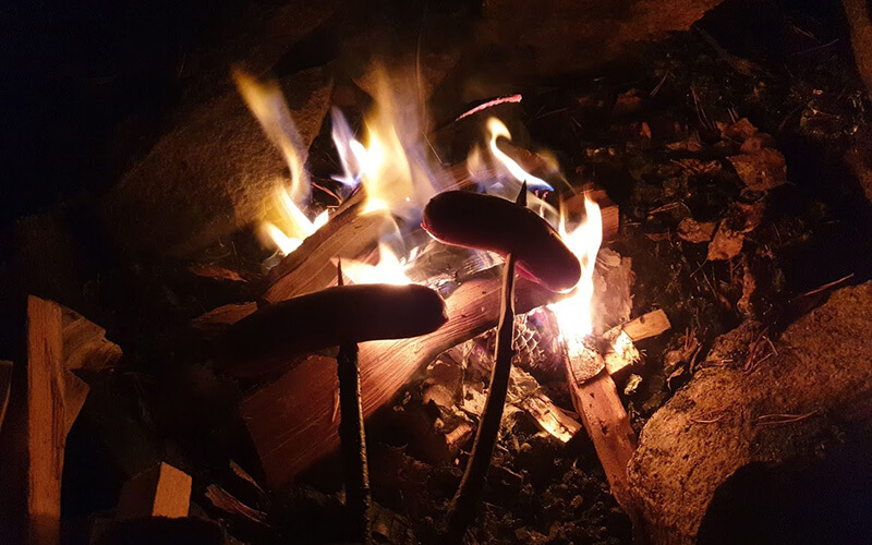 Zwei Würstchen auf Stöcken über einem offenen Lagerfeuer.