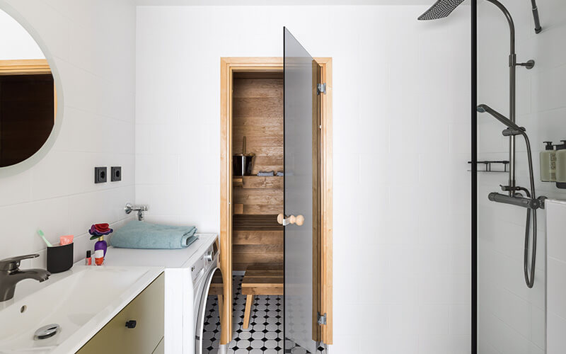 Badezimmer mit Waschbecken, Waschmaschine, einer Duschkabine mit Regenduschkopf und Blick durhc eine offene Glastür in eine Sauna.