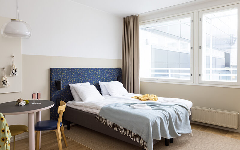 Schlafzimmer mit Doppelbett, blau gemustertem Kopfteil, zwei Stühlen an rundem Tisch und großem Fenster mit Vorhang.