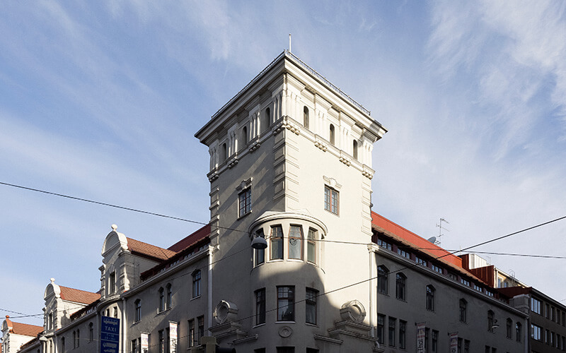 Eckgebäude mit Turm und roten Dächern unter blauem Himmel.