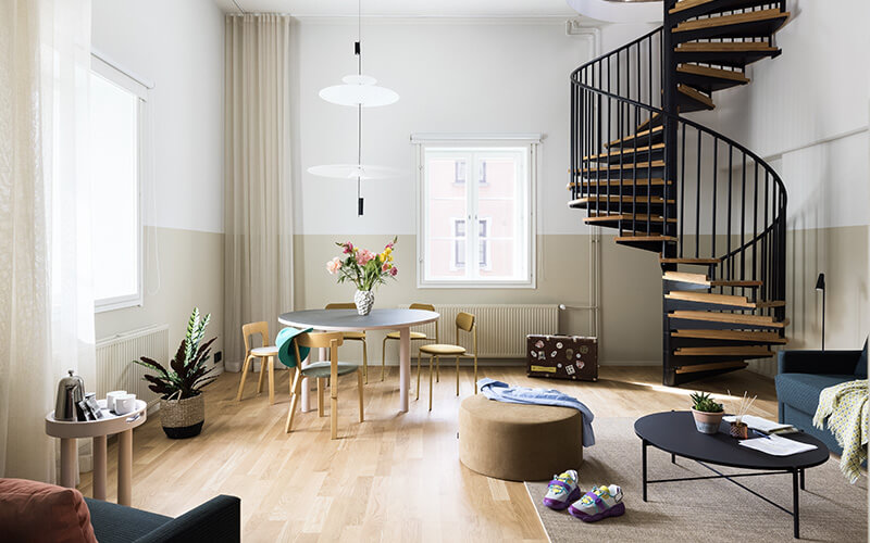 Helles Wohnzimmer mit runder Esstischgruppe und Sitzmöbeln auf hellem Holzboden. Eine Wendeltreppe führt nach oben.