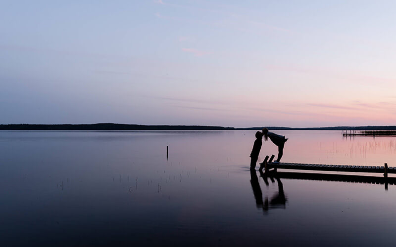 Zwei Personen auf einem Steg über ruhigem Wasser bei Sonnenuntergang, eine Person beugt sich vor, um die andere zu küssen.
