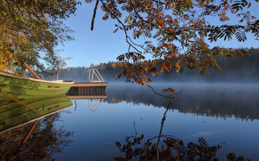 Stillgelegtes Boot am Ufer eines nebligen Sees mit Steg und herbstlichen Zweigen im Vordergrund.