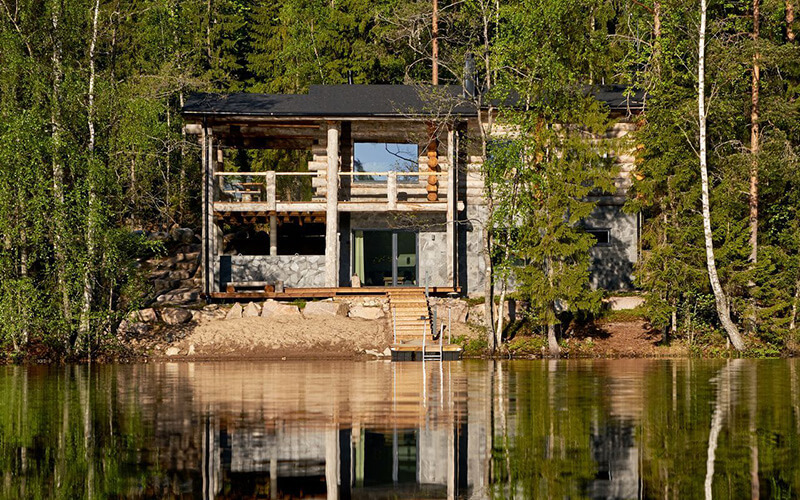Holzhaus mit großer Glasfront und Balkon am Ufer eines ruhigen Sees umgeben von Bäumen.