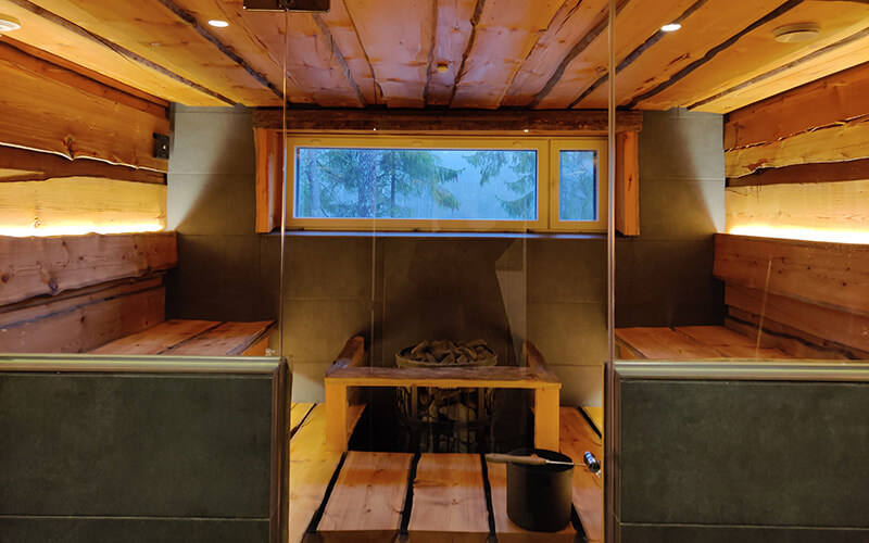 Innenraum einer finnischen Sauna mit Holzbänken, einem Ofen mit Steinen und einem schmalen Fenster mit Blick auf Bäume.