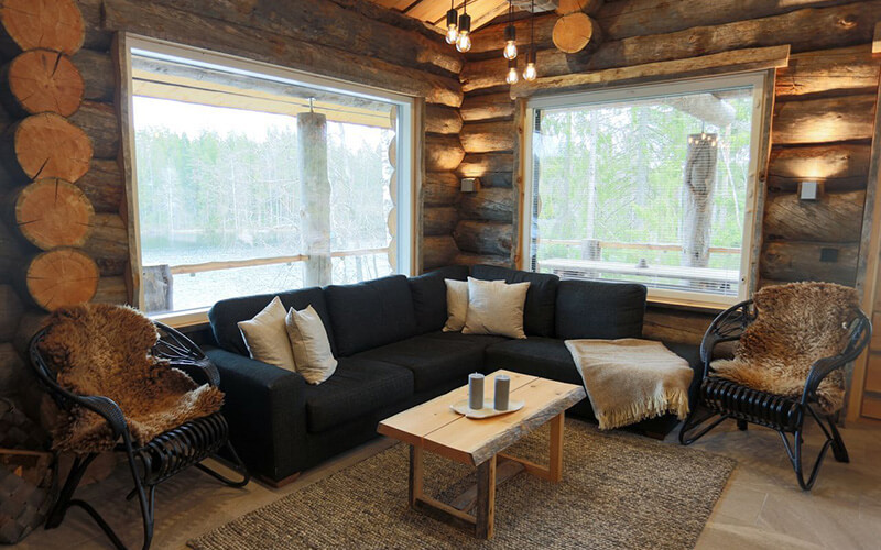 Gemütliche Sofaecke mit dunklem Ecksofa, zwei Korbsesseln mit Fellbezügen, Holztisch und großen Fenstern in einer Blockhütte.