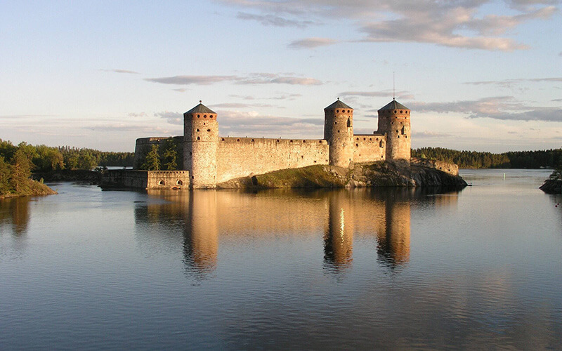 Steinerne Burg mit drei runden Türmen auf einer Insel im ruhigen Wasser unter bewölktem Himmel.