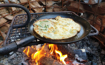 Gusseiserne Pfannne mit Pfannkuchen über Feuer mit Holzscheiten im Hintergrund.