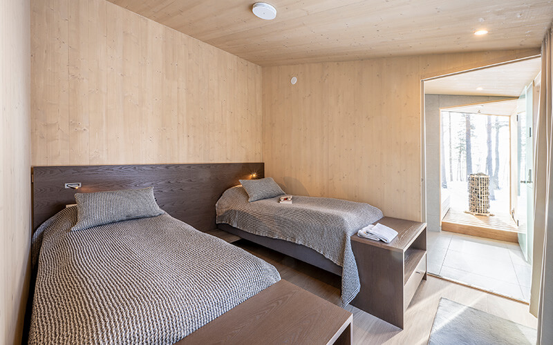 Kinderzimmer mit zwei Einzelbetten, grauen Decken und Holzwänden, Blick in einen hellen Flur und Sauna mit Glasfront.