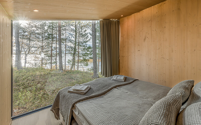 Schlafzimmer mit Holzwänden, Doppelbett mit grauer Bettwäsche und großen Fenstern mit Blick auf Bäume und Wasser.