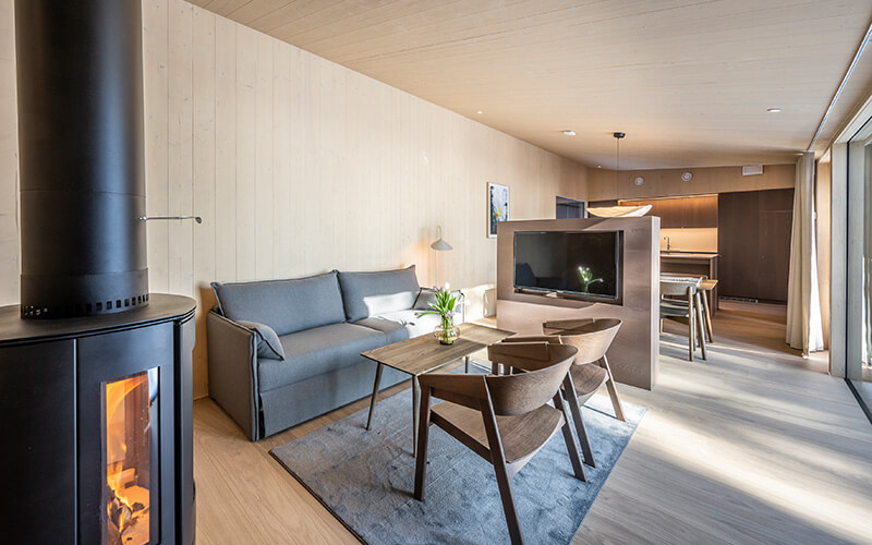 Moderner Wohnbereich mit grauem Sofa, zwei Holzstühlen, Kaminofen und offener Küche im Hintergrund.