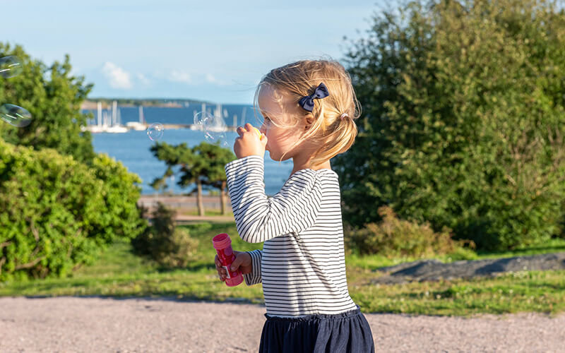 Ein Mädchen mit gestreiftem Oberteil und dunklem Rock pustet Seifenblasen in einer Parklandschaft mit Wasser und Brücke im Hintergrund.