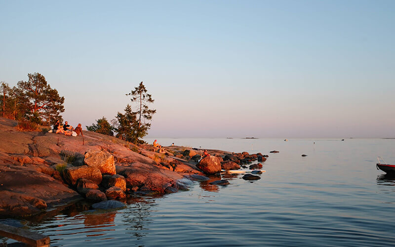 Felsige Küste mit wenigen Bäumen und Menschen am Ufer eines ruhigen Gewässers bei Sonnenuntergang.
