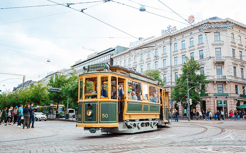 Historische Straßenbahn mit Passagieren fährt auf einer kurvigen Straße in einer belebten Innenstadt mit Altbauten und Fußgängern.