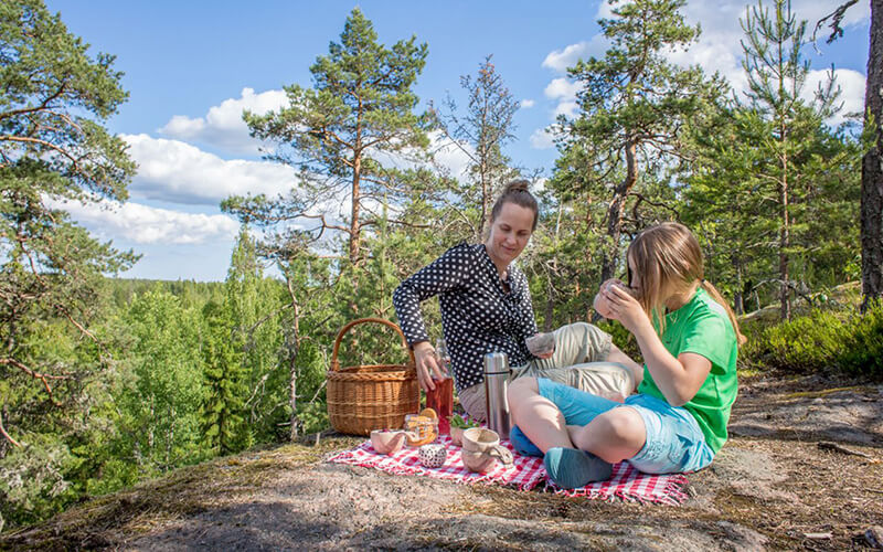 Zwei Personen sitzen auf einer karierten Decke auf einem Felsen im Wald und machen Picknick mit Korb und Thermoskanne.