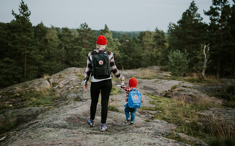 Erwachsene und Kind mit roten Mützen und Rucksäcken wandern auf felsigem Gelände in bewaldeter Landschaft.