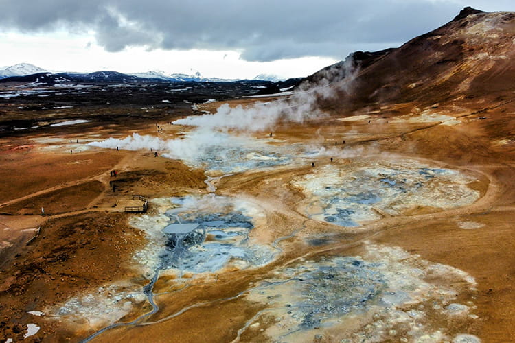 Landschaft mit dampfenden geothermalen Quellen und felsigem Gelände vor schneebedeckten Bergen unter bewölktem Himmel