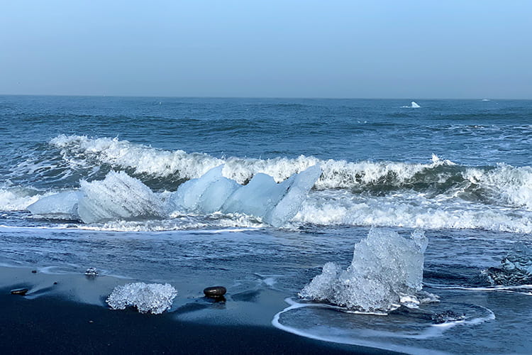 Eisschollen am schwarzen Sandstrand mit Wellen im Meer unter klarem Himmel