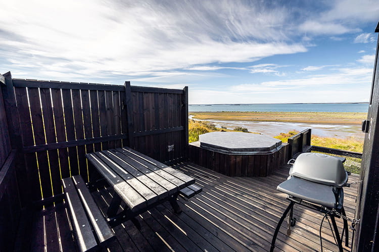 Holzterrasse mit Eck-Whirlpool, Holztisch mit Bank und Grill vor Blick auf Strand und Meer